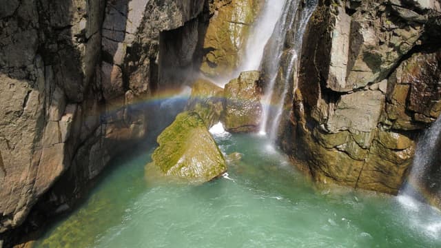A scenic view of a waterfall flowing over rugged, brown rock formations into a clear, green-blue pool, with a faint rainbow arching through the spray near a moss-covered boulder.