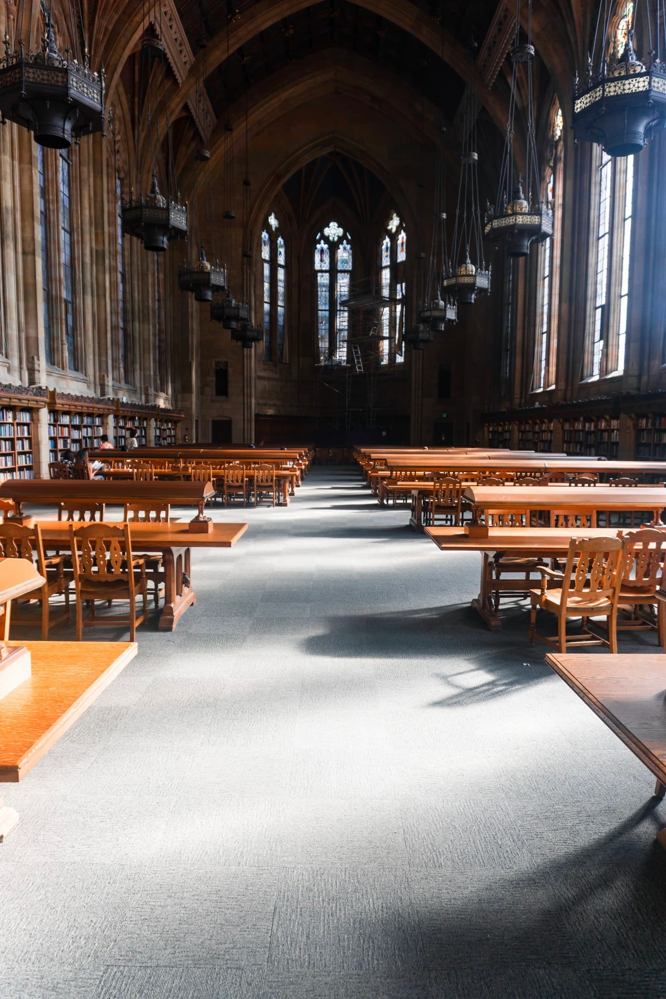 A large library hall with rows of wooden tables, bookshelves, and sunlight streaming through tall windows