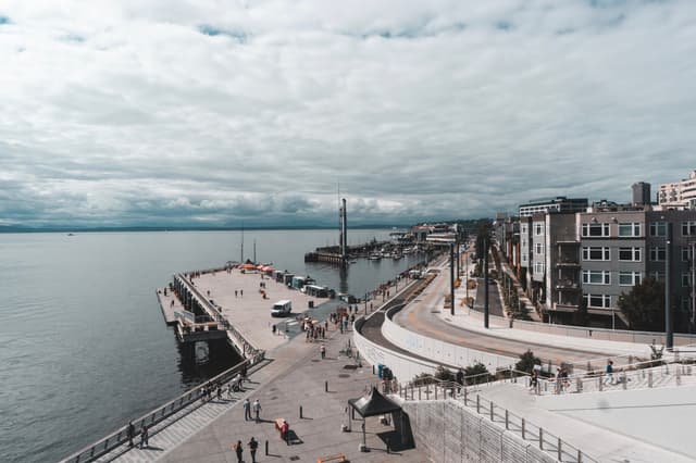A paved waterfront walkway features a long pier and adjacent buildings