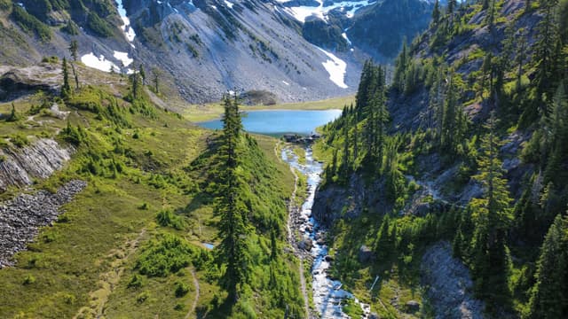 A high-angle shot captures a vibrant green valley with a rocky stream leading to a serene blue lake, framed by steep mountain slopes with patches of snow and scattered evergreen trees.