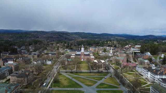 A high-angle shot overlooking a town featuring a central grassy area with intersecting paths, a white fence, and a building with a tall steeple, set against a backdrop of rolling hills under a cloudy sky.