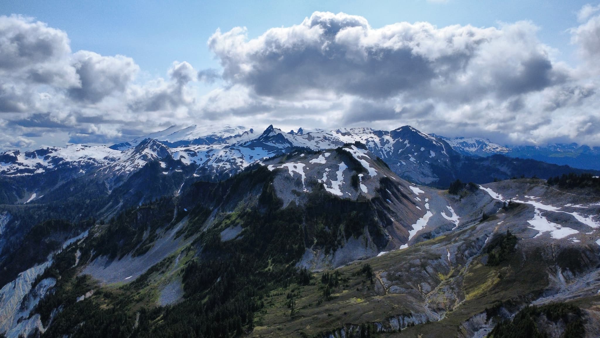 A panoramic view of a rugged mountain range featuring snow-dusted peaks, rocky slopes with sparse vegetation, and a bright blue sky filled with fluffy white clouds.