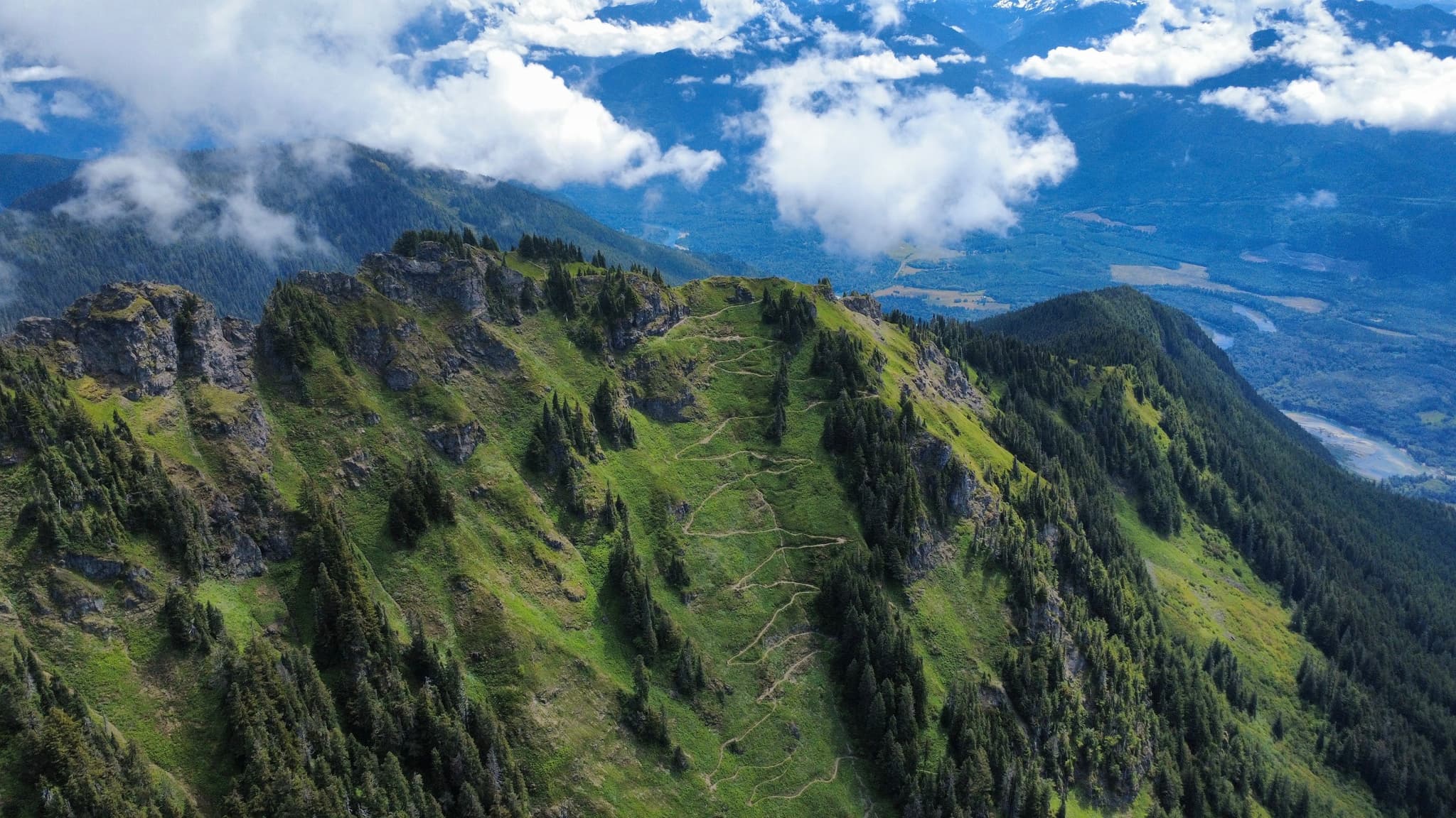 An aerial view captures a lush green mountain ridge with scattered trees, partially obscured by white clouds against a bright blue sky.