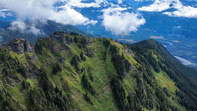 An aerial view captures a lush green mountain ridge with scattered trees, partially obscured by white clouds against a bright blue sky.