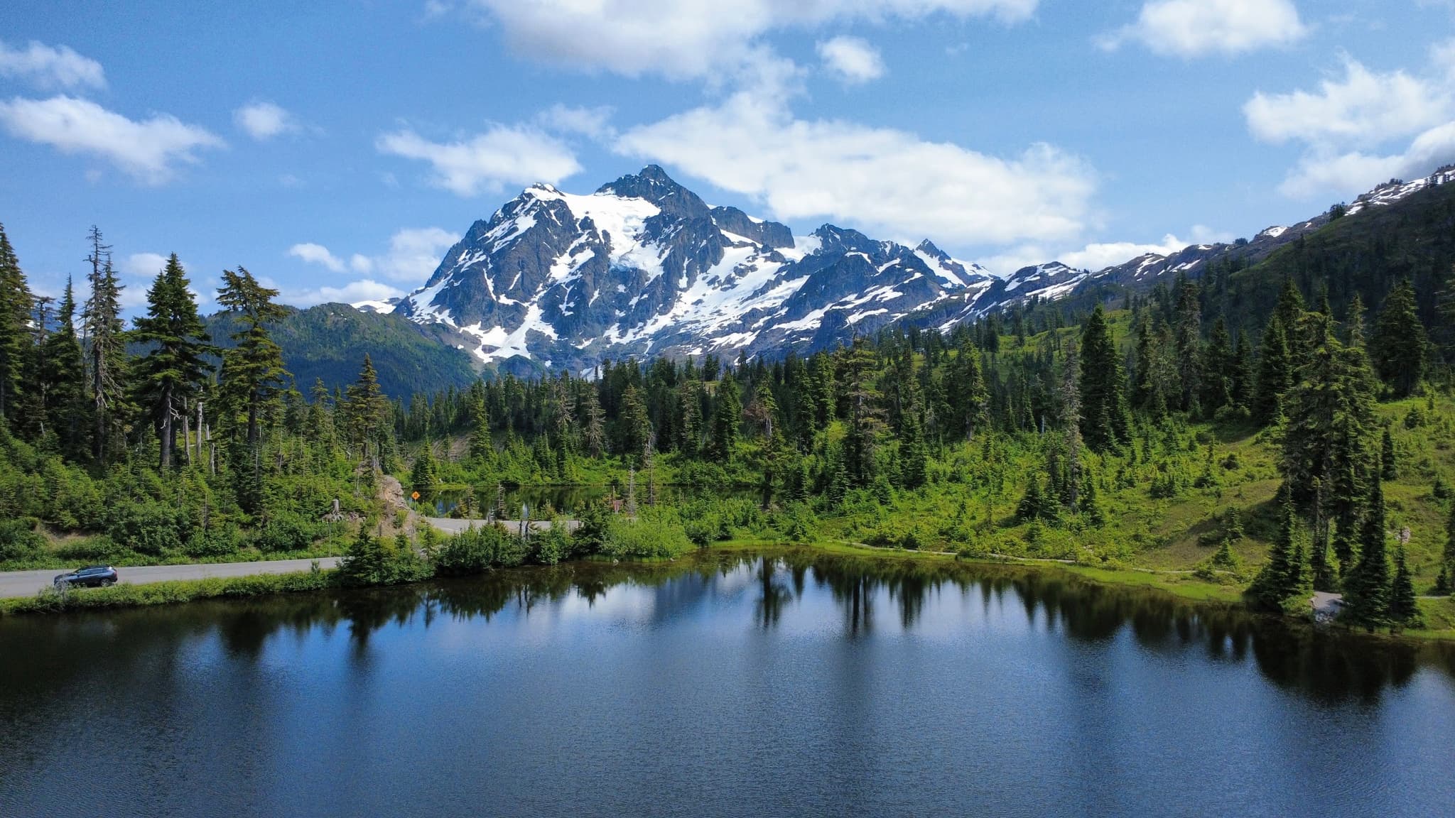 A tranquil landscape featuring a calm lake that mirrors a majestic snow-covered mountain and a dense forest of evergreen trees, set against a bright blue sky with scattered clouds.