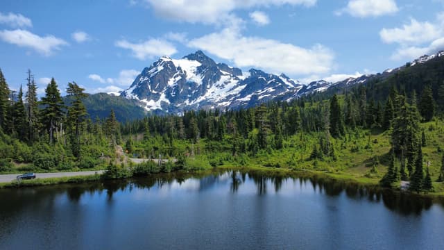 A tranquil landscape featuring a calm lake that mirrors a majestic snow-covered mountain and a dense forest of evergreen trees, set against a bright blue sky with scattered clouds.